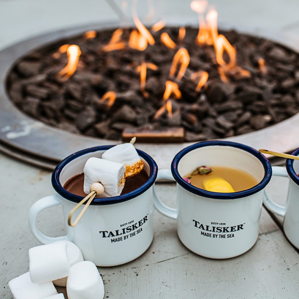 Three Talisker mugs with marshmallows near a GlammFire gas fire pit with vibrant flames, creating a cozy outdoor setting.