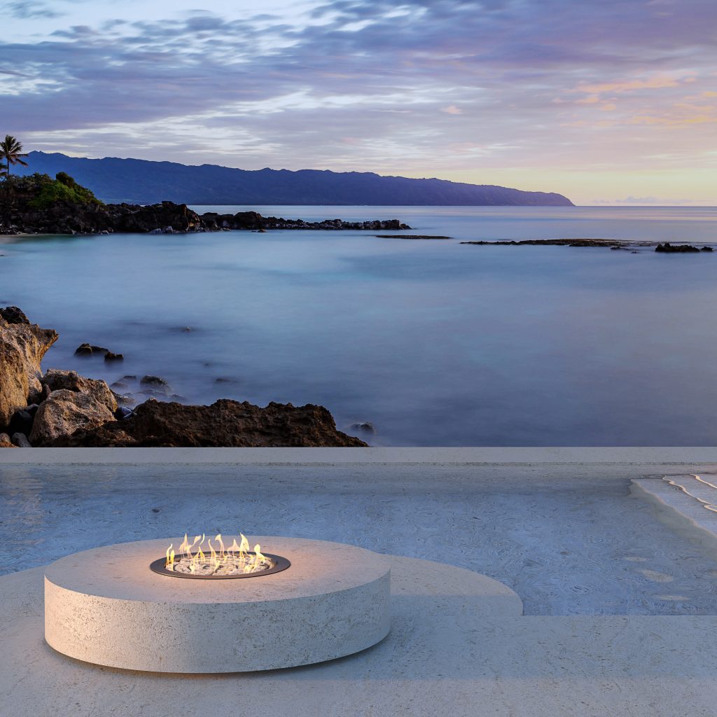 Round gas fire pit by GlammFire, placed in an outdoor setting with a serene coastal view, ideal for outdoor heating and modern design.