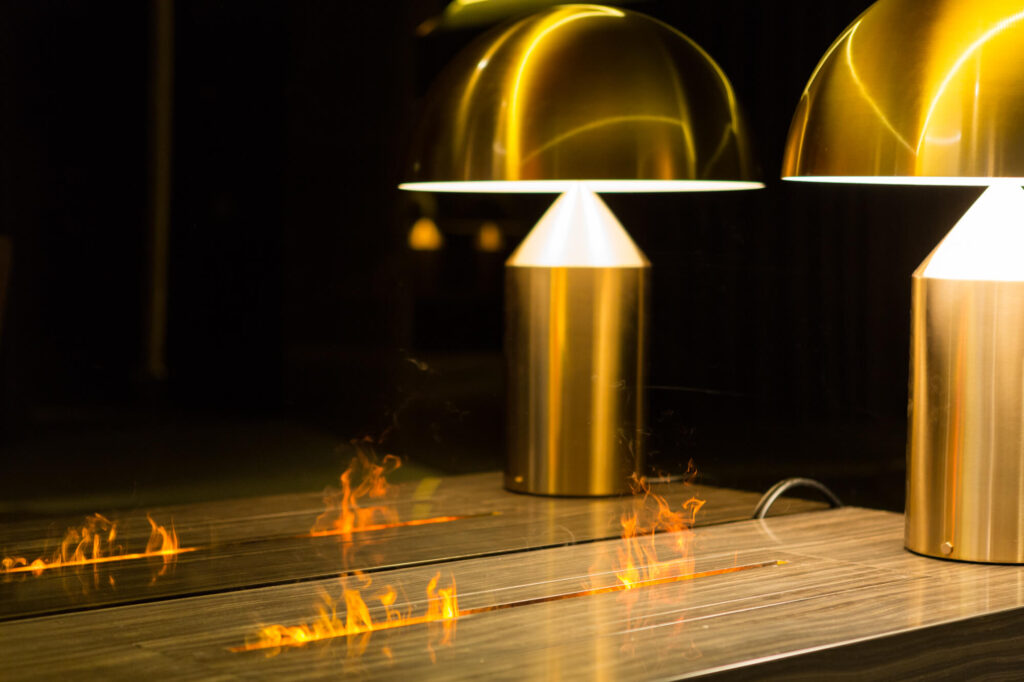 lose-up of GlammFire Gema fireplace flames reflected on a wooden surface beside modern golden dome lamps in a contemporary interior setting
