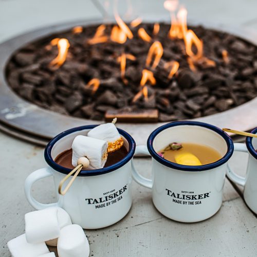 Three Talisker mugs with marshmallows near a GlammFire gas fire pit with vibrant flames, creating a cozy outdoor setting.