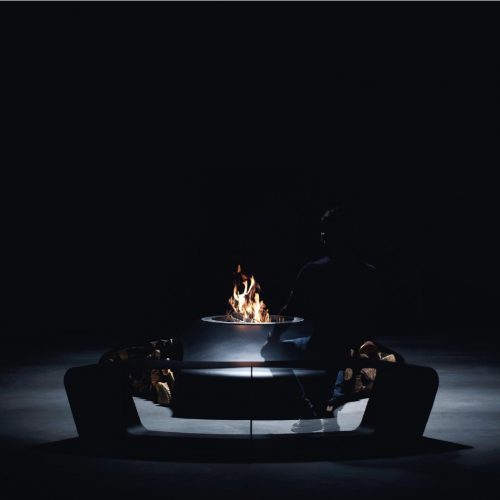 Vaudeville fire pit in a minimalistic dark setting with a person sitting nearby.