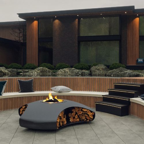 The Vaudeville fire pit, featuring a sleek design with integrated wood storage, complements the modern architecture and outdoor seating area.