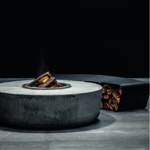 Industrial-style Zarzuela Fire Pit by GlammFire with a concrete body, burning wood logs, and a sleek firewood storage bench.